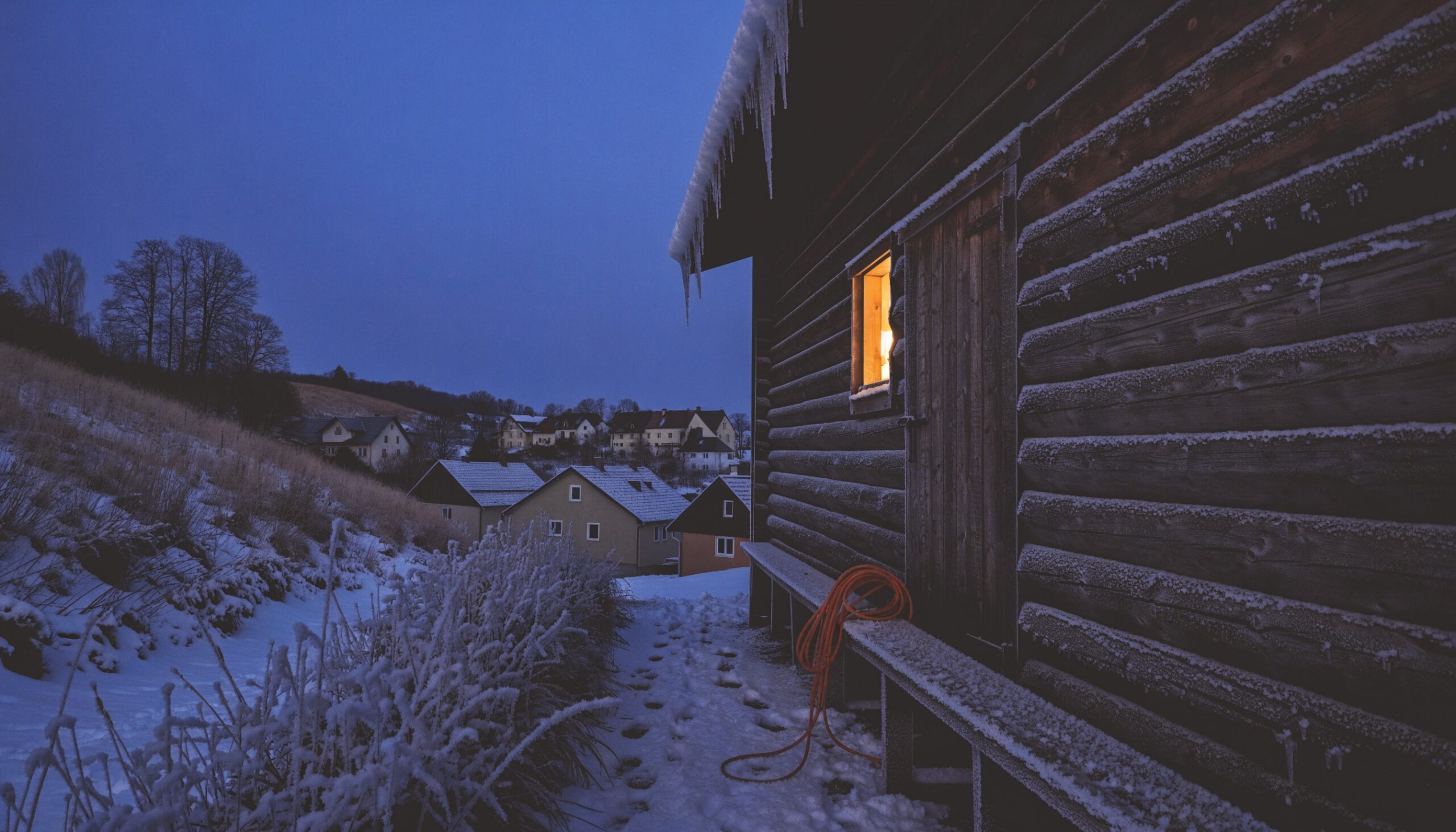 Bauernhof in der Winterdämmerung mit beleuchtetem Stallfenster und Verlängerungskabel – Zapfwellengenerator für den Stromausfall